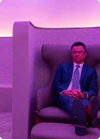 A man in a blue suit sits with his eyes closed and hands clasped in a high-backed chair within a private seating nook bathed in pink and purple light.
